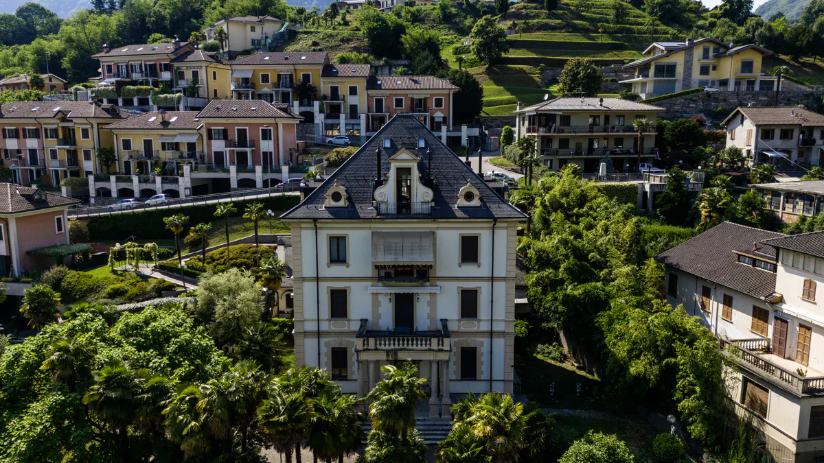 Aerial view of a grand, light-colored villa surrounded by lush trees and a hillside neighborhood of colorful homes.