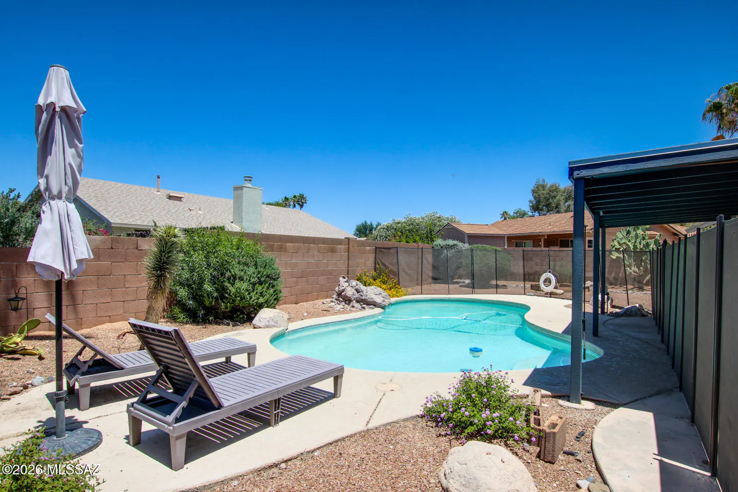 Backyard with a blue pool, two gray lounge chairs, and a closed white umbrella. A brick wall surrounds the yard.