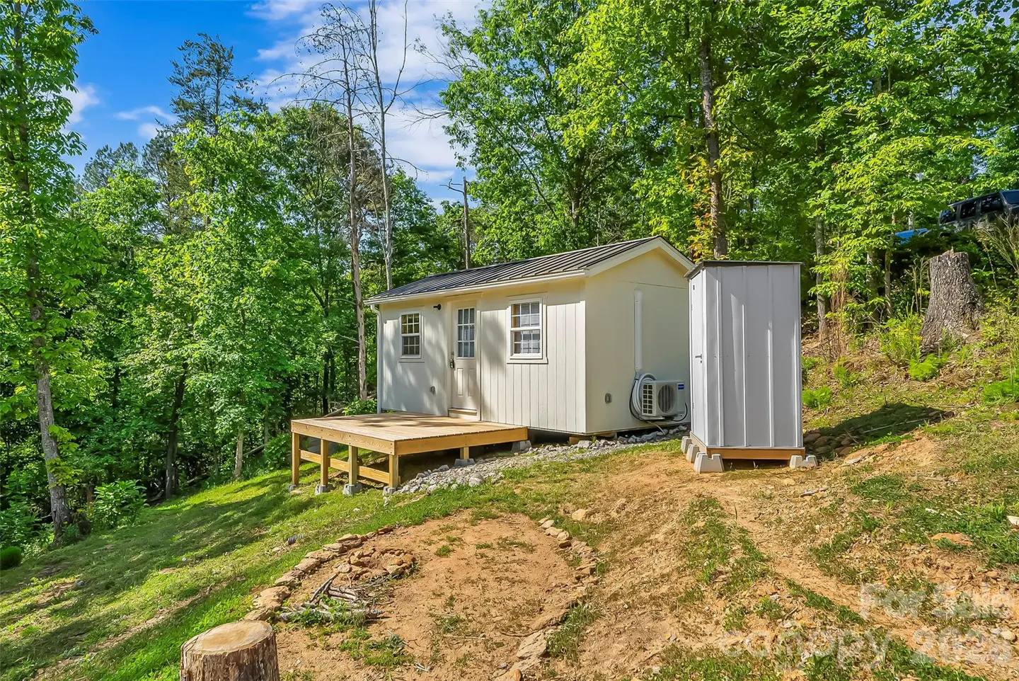 Exterior view of a small, cream-colored cabin with a wooden deck, surrounded by lush green trees on a sunny day.