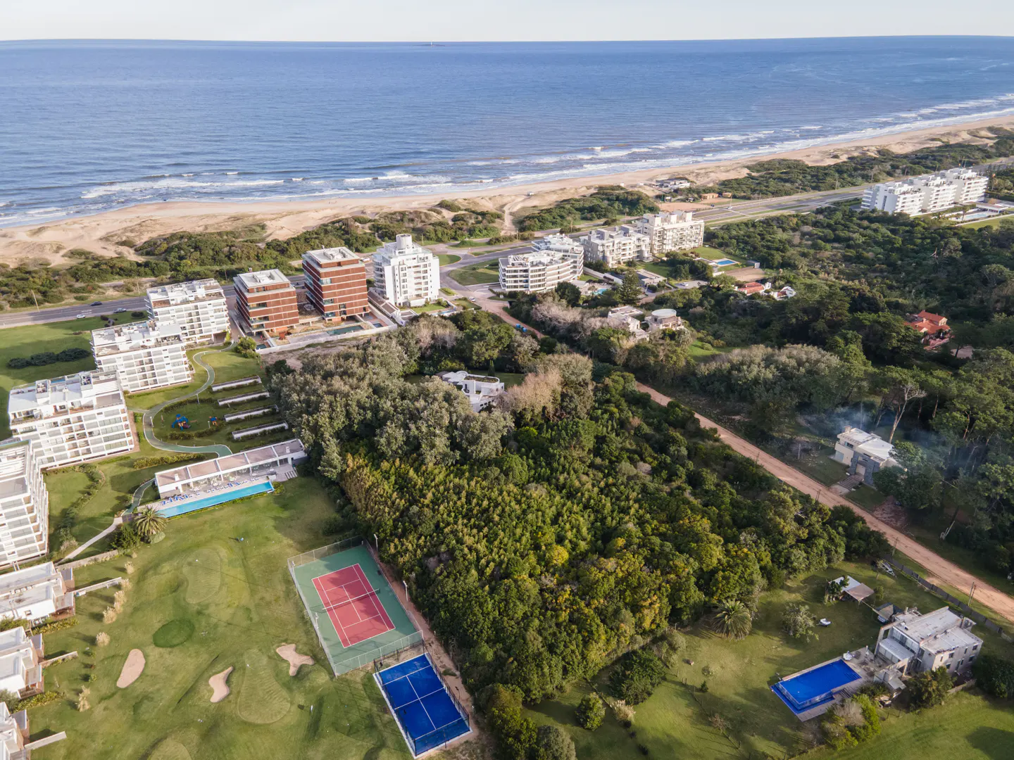 Aerial view of beachfront property with condos, golf course, tennis courts, and swimming pools, set against a blue ocean backdrop.
