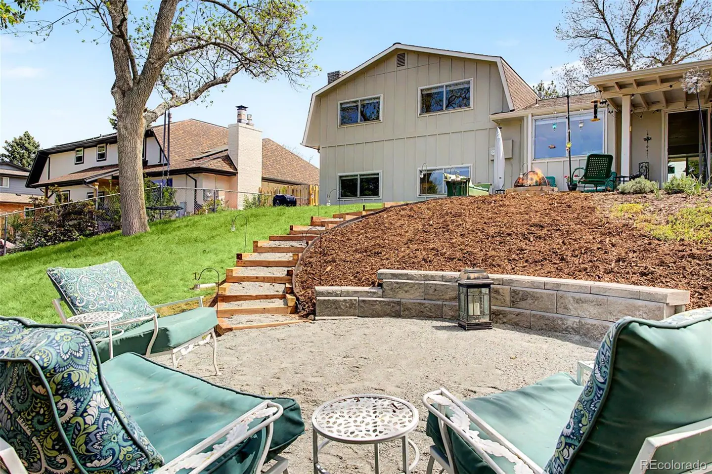 Backyard patio with teal cushioned chairs, small tables, and stone retaining wall leading to a beige house with a brown roof.