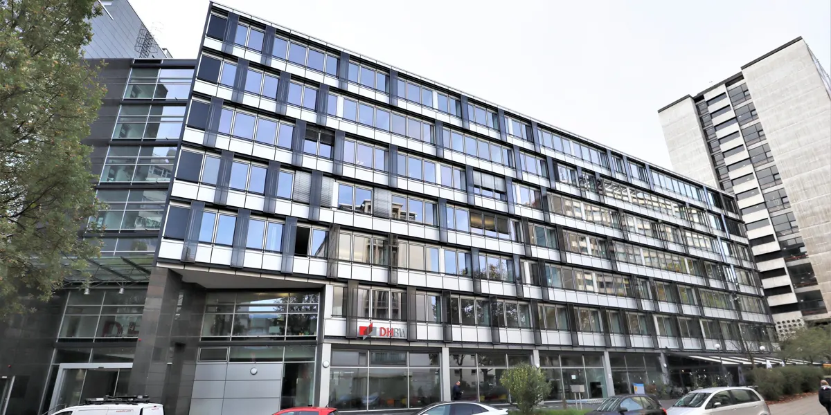 Exterior view of a modern, multi-story office building with a glass facade and black window frames. Cars are parked in front.