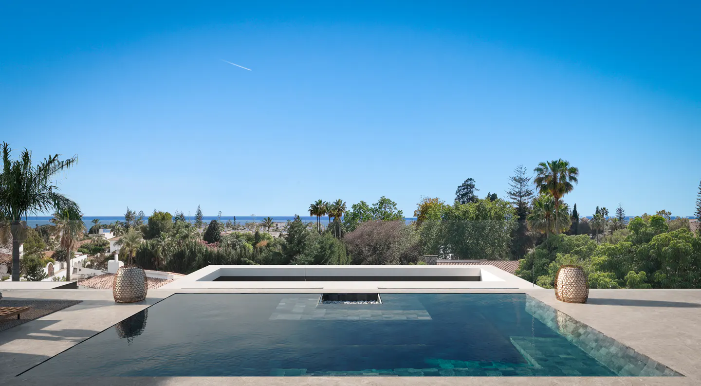 Infinity pool on a modern terrace overlooking lush trees and the distant ocean under a clear blue sky.
