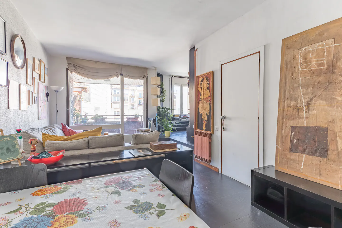 Bright living room with floral tablecloth, gray sofa, and large windows. Artwork adorns the walls, and a black console table adds a modern touch.