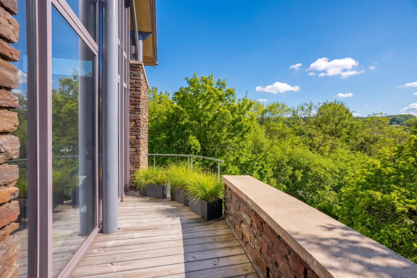 A modern balcony with stone walls, a wood floor, and metal railings overlooks lush green trees under a blue sky. Tall windows reflect the scenery.