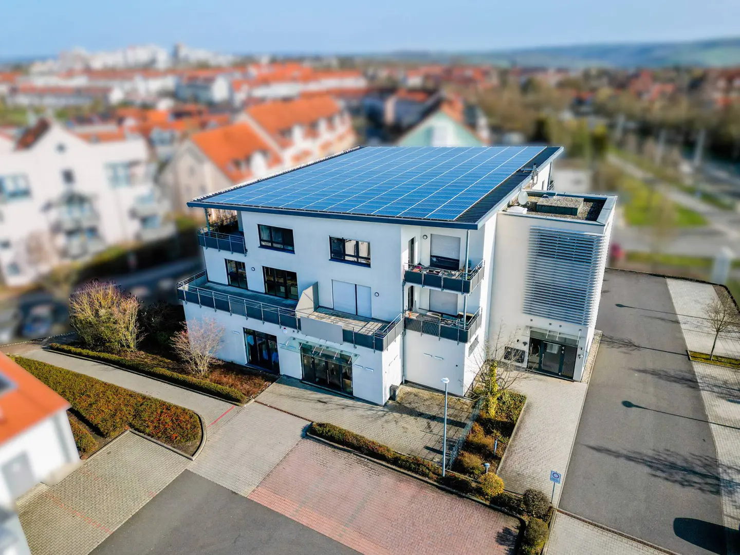 Aerial view of a modern, white apartment building with solar panels on the roof, balconies, and a landscaped yard.