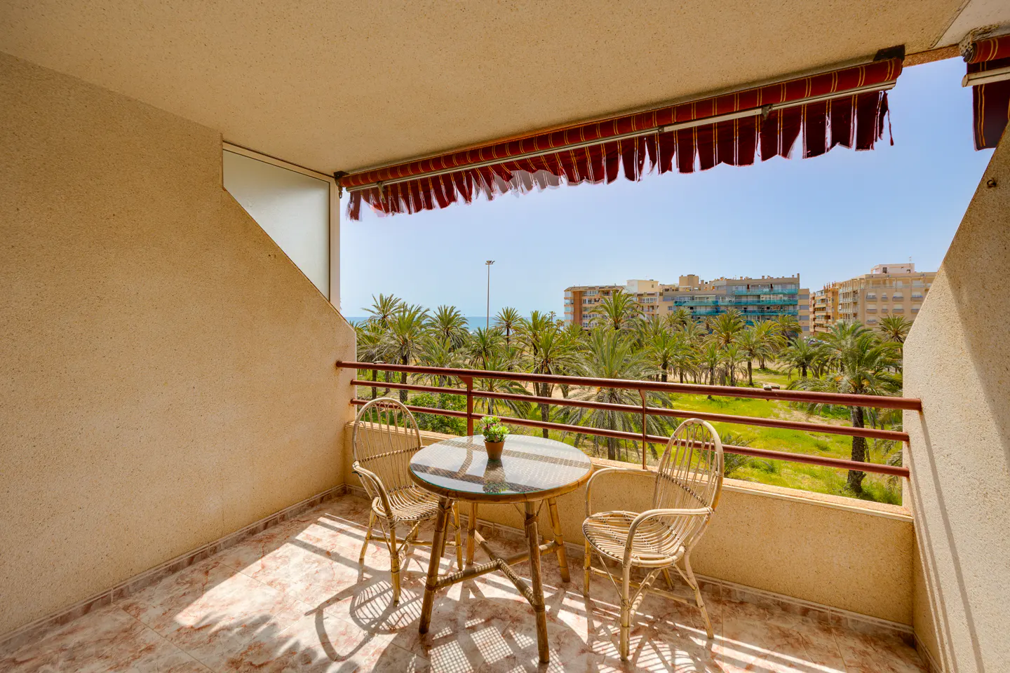 Balcony view with a round table, two wicker chairs, and a small plant. Palm trees and buildings are visible in the background. Red and white awning.