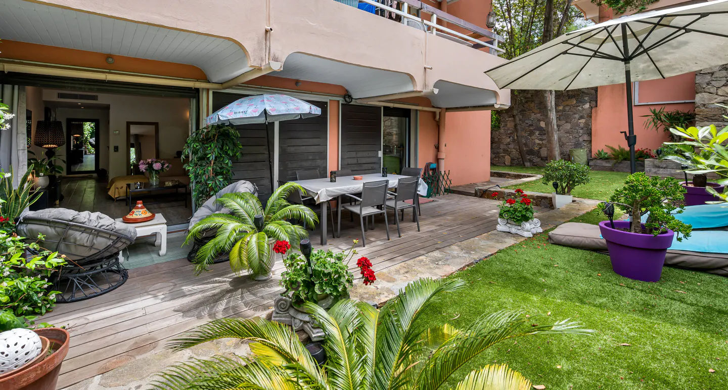 Outdoor patio with a table, chairs, and lounge seating. Lush greenery surrounds the space, with a building in the background.