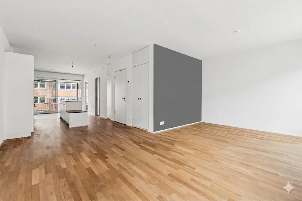 Bright, empty room with hardwood floors, white walls, and a gray accent wall. Balcony visible through large windows.