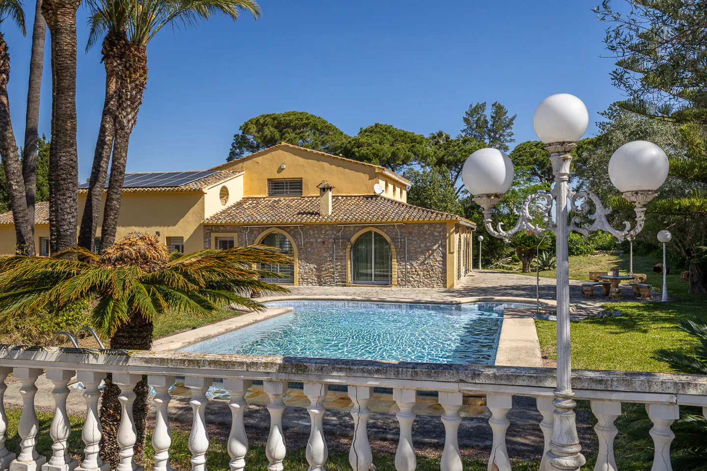 A yellow house with a pool and palm trees on a sunny day. A white fence is in the foreground.