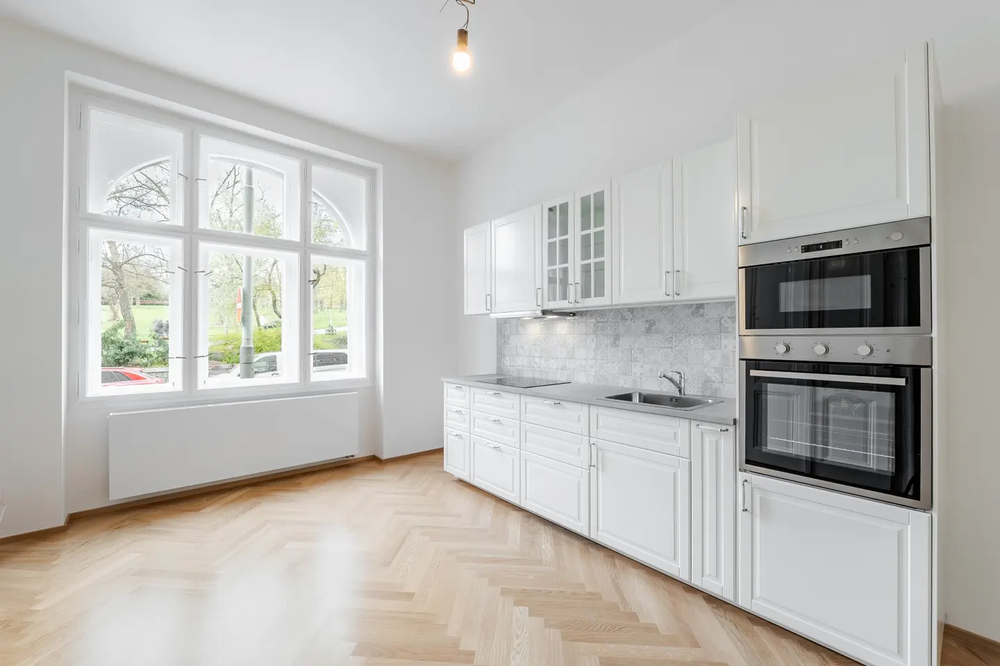 Bright, white kitchen with herringbone wood floors, stainless steel oven, and large window overlooking a park.