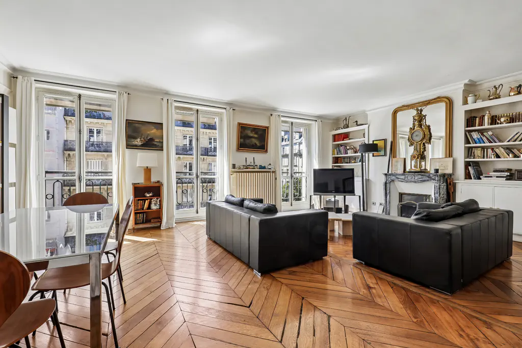 Bright living room with herringbone wood floors, black sofas, dining table, and windows overlooking a city street.