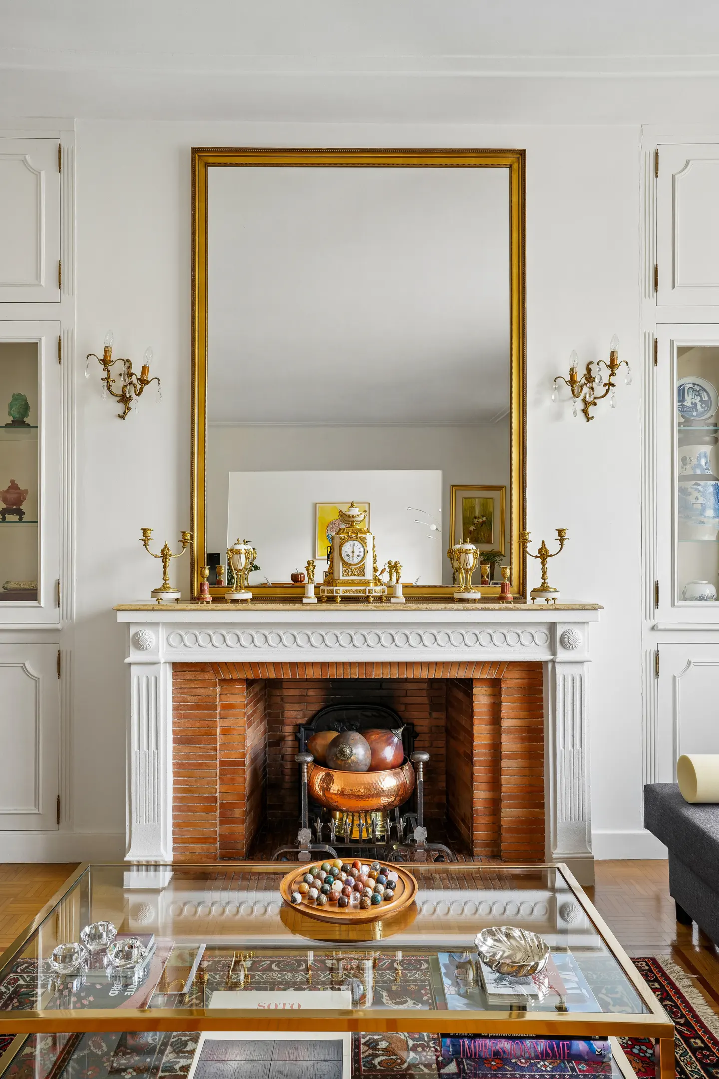A white fireplace with a brick interior, topped by a large gold-framed mirror, flanked by built-in cabinets and wall sconces.
