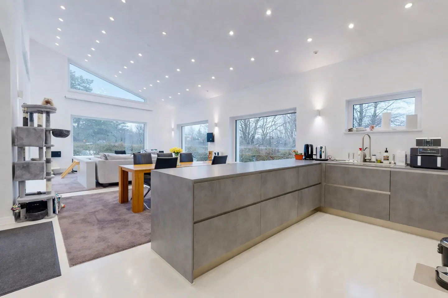 Bright, open-concept kitchen and living area with white walls, gray cabinets, wood table, and large windows overlooking trees.