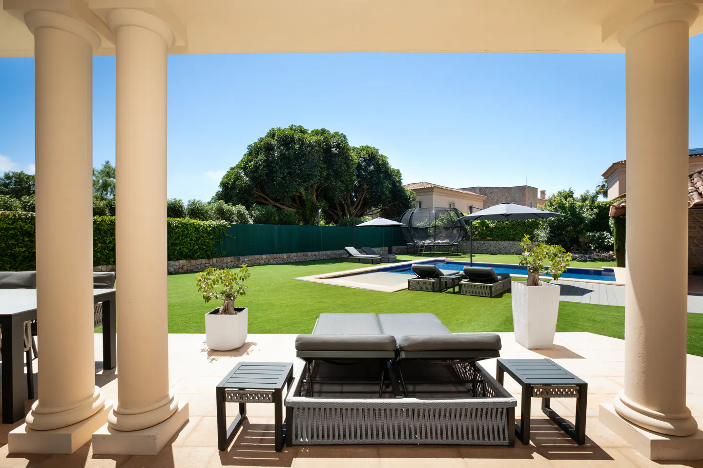 View of a backyard with a pool and lounge chairs, framed by white columns. Green lawn, trees, and blue sky in the background.