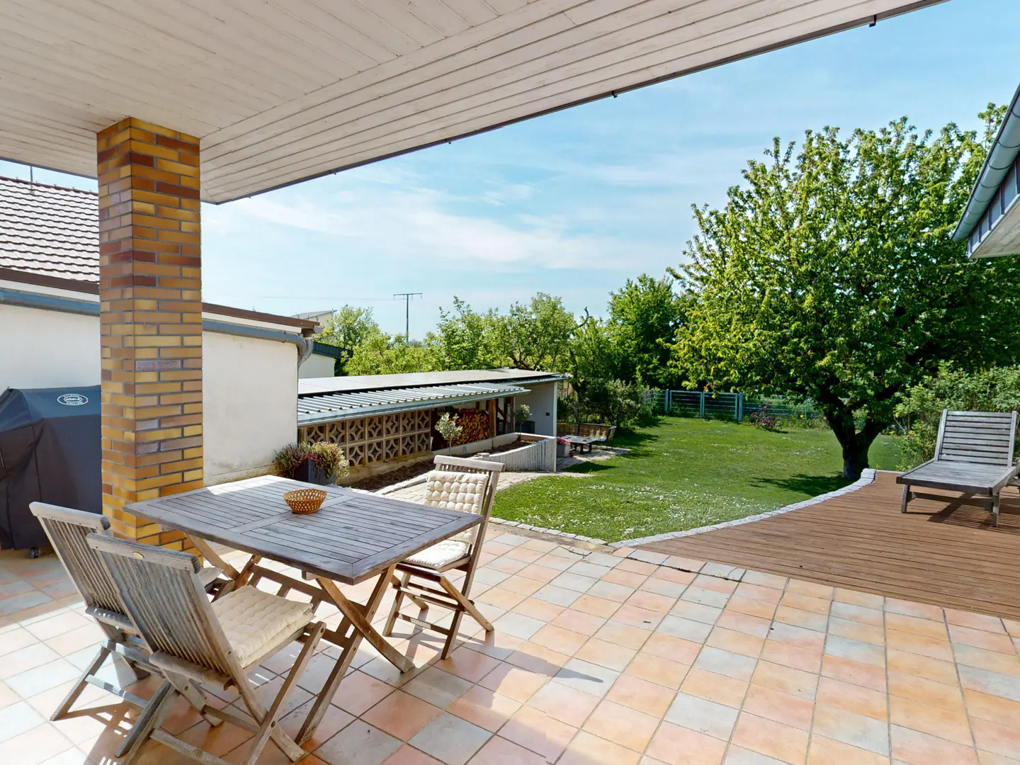 Outdoor patio with a wooden table and chairs on a tiled floor, leading to a green lawn with trees and a wooden deck.