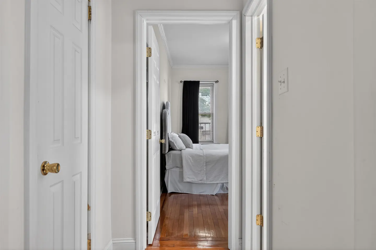 View through a white hallway into a bedroom with a bed, white bedding, and black curtains against a window.