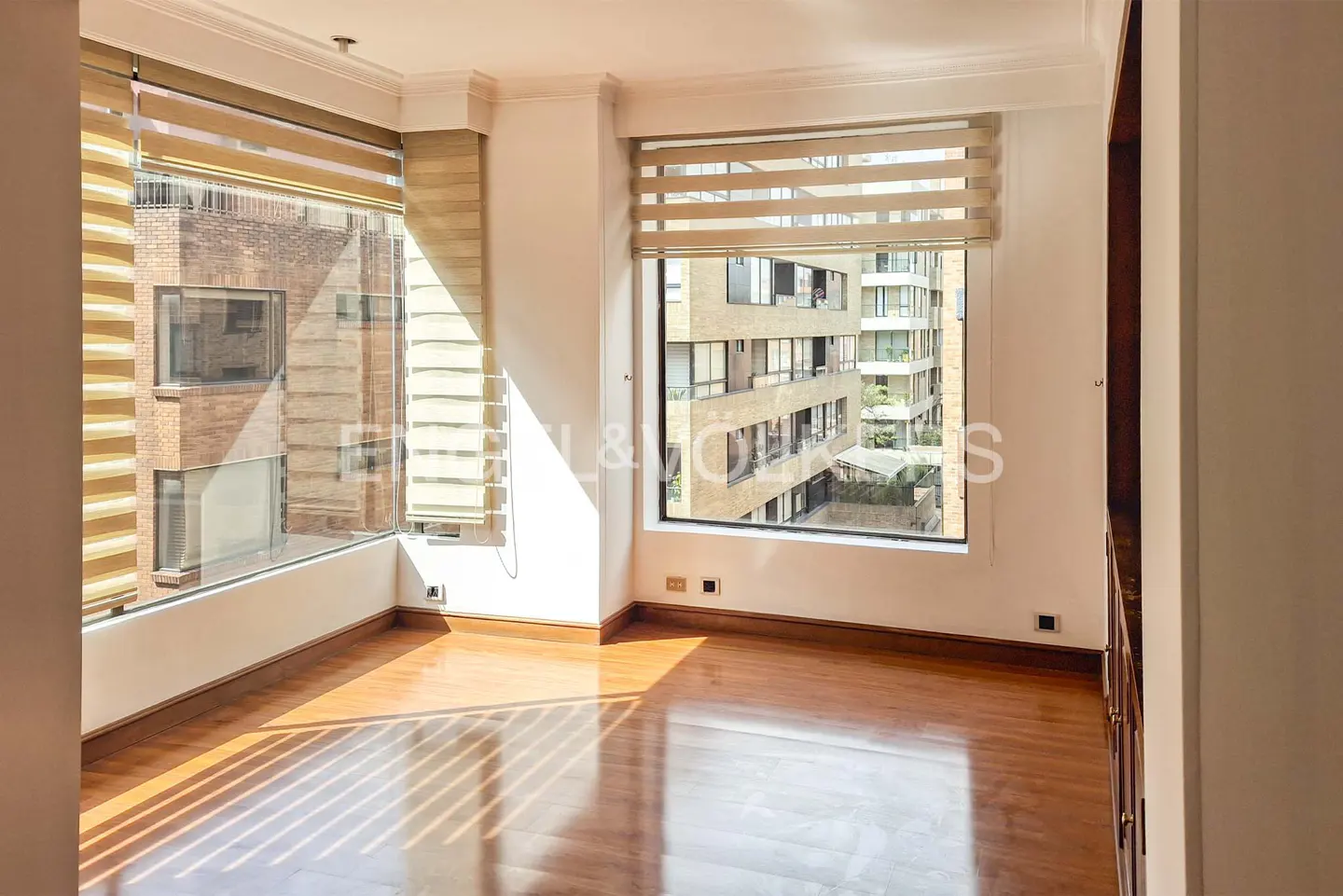 Bright room with hardwood floors, large windows featuring tan blinds, and a view of neighboring apartment buildings.