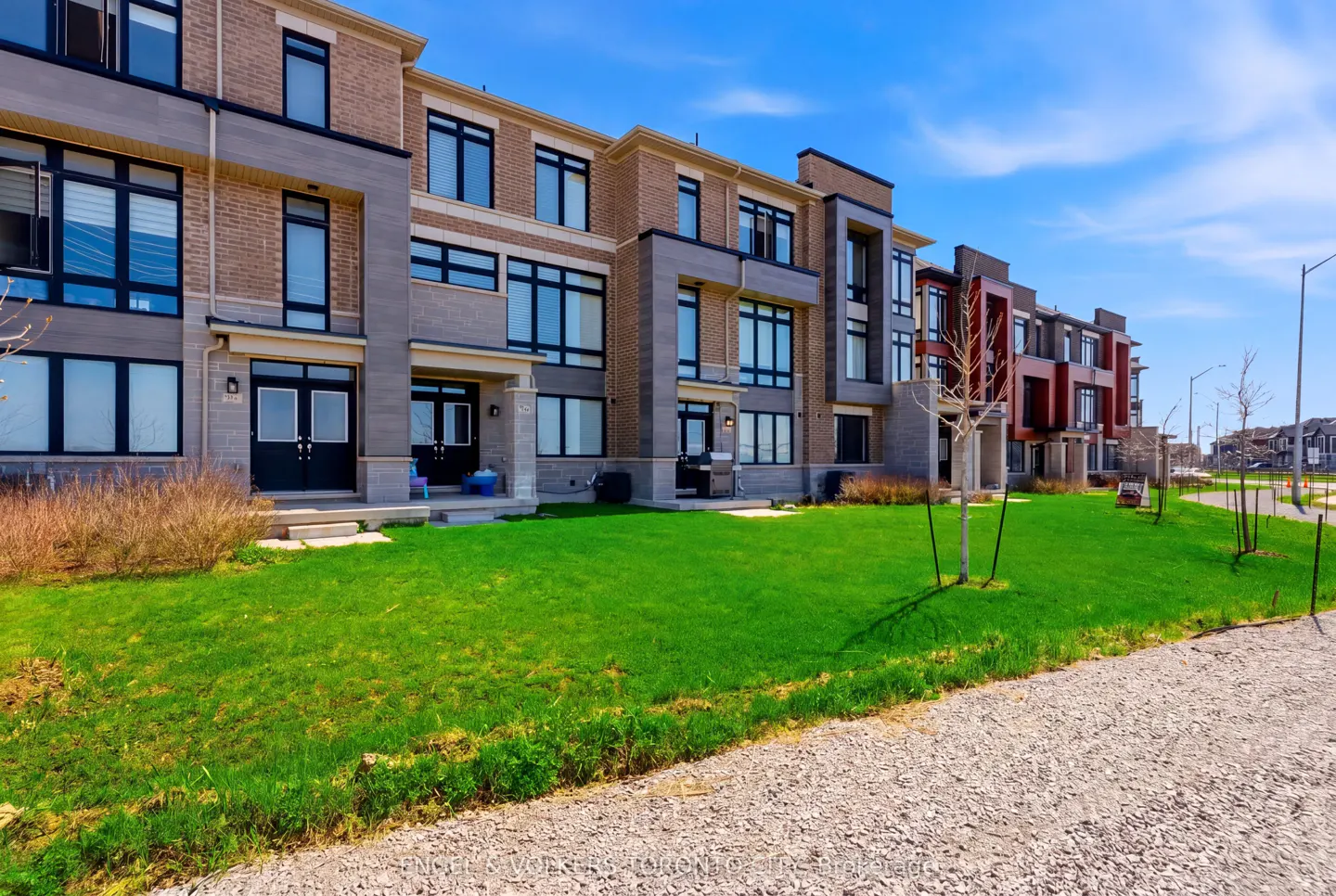 Row of modern townhouses with brick and gray stone exteriors, black windows, and green lawns under a bright blue sky.
