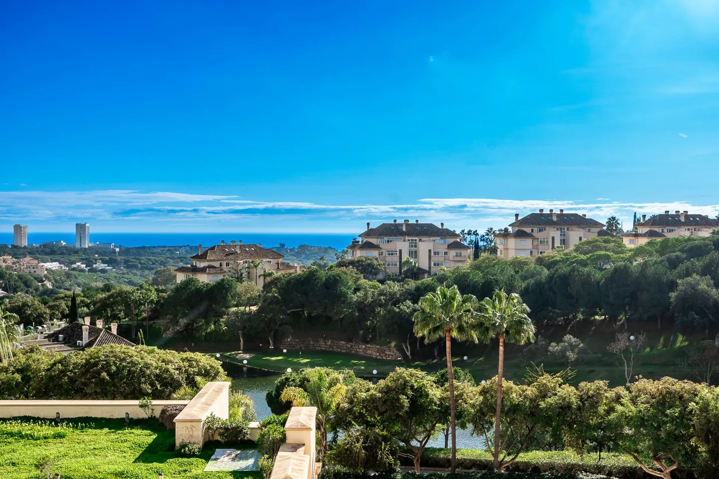 Scenic view of luxury homes nestled in lush greenery, with the ocean and city skyline in the background under a clear blue sky.