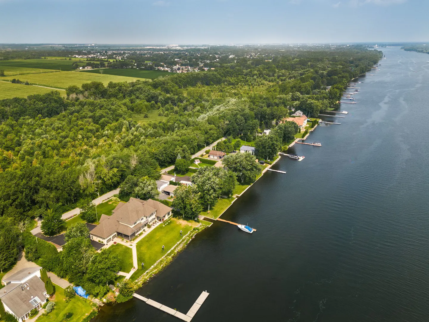 Aerial view of waterfront homes with docks on a river, surrounded by lush green trees and fields under a blue sky.