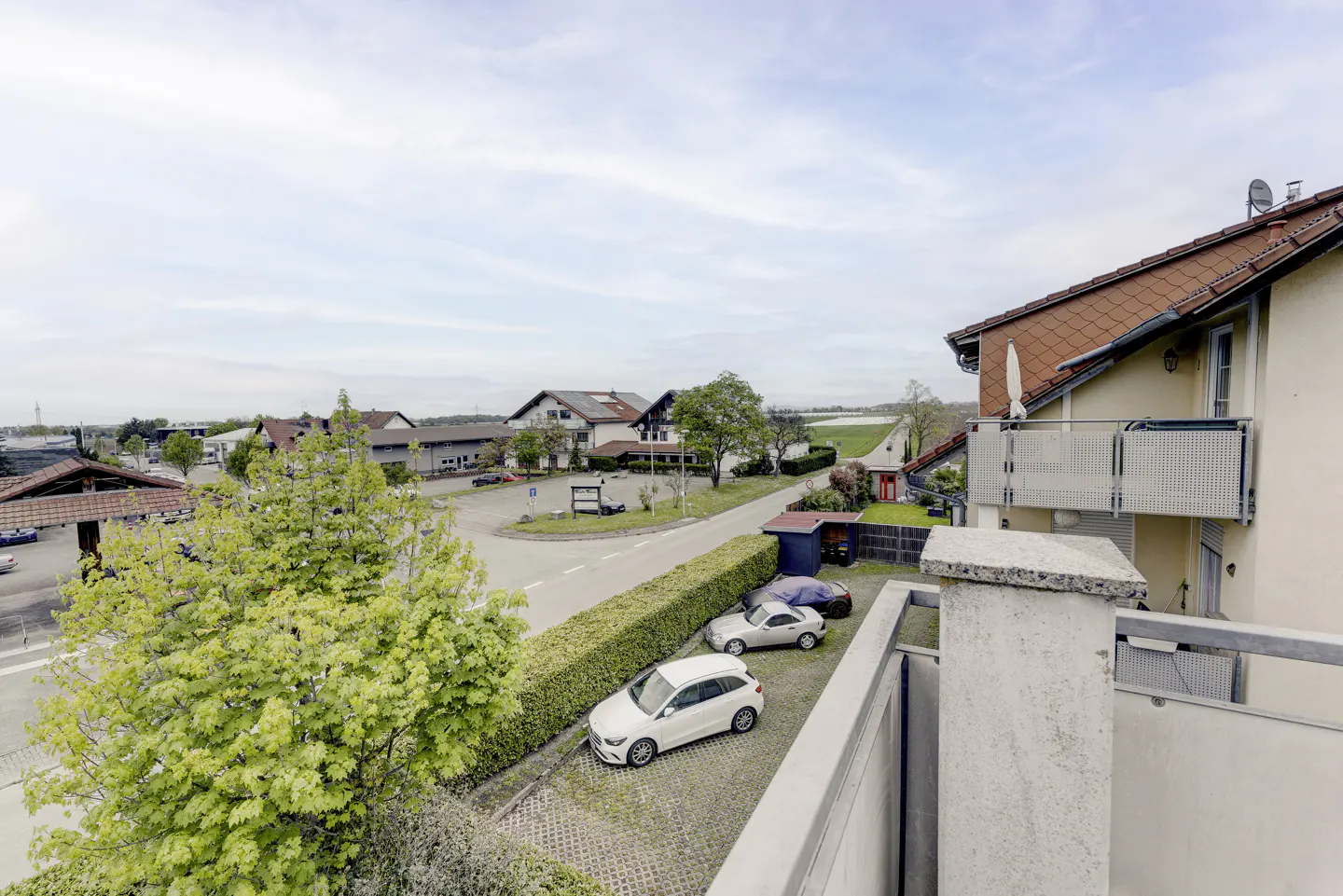 View from a balcony overlooking a street with parked cars, houses, and green trees under a cloudy sky.
