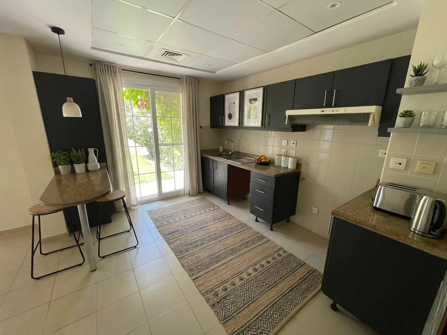 A bright kitchen with black cabinets, a marble countertop, and a patterned rug. A glass door leads to a garden.