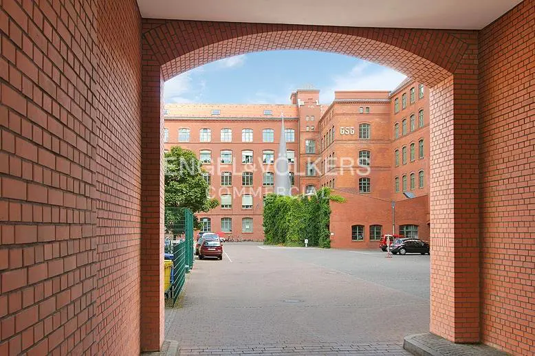 View of a red brick building through an arched brick entryway. Cars are parked in the lot. The sky is blue with some clouds.