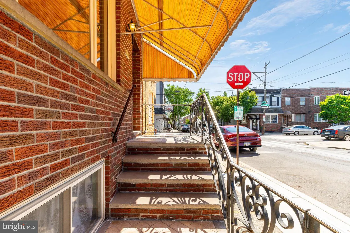 Brick steps with black railings lead to a house with a yellow awning. A stop sign and parked cars are visible on the street.