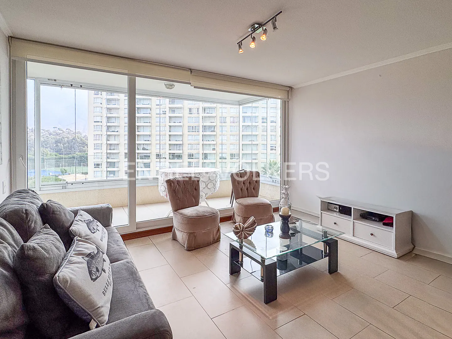 Living room with a gray sofa, glass coffee table, and two chairs facing a balcony with a city view.