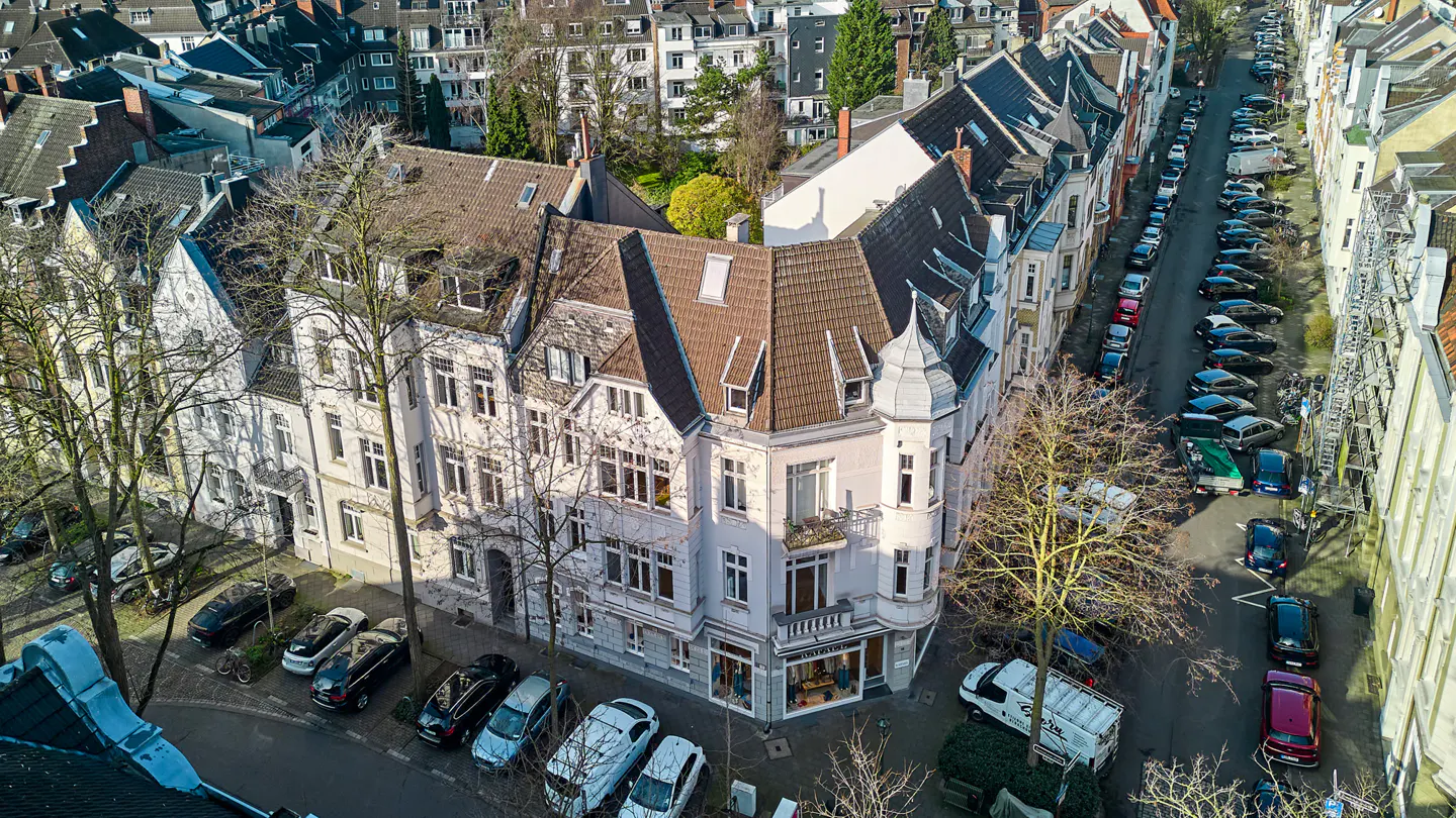 Aerial view of a white corner building with a brown roof, surrounded by parked cars on a narrow city street.