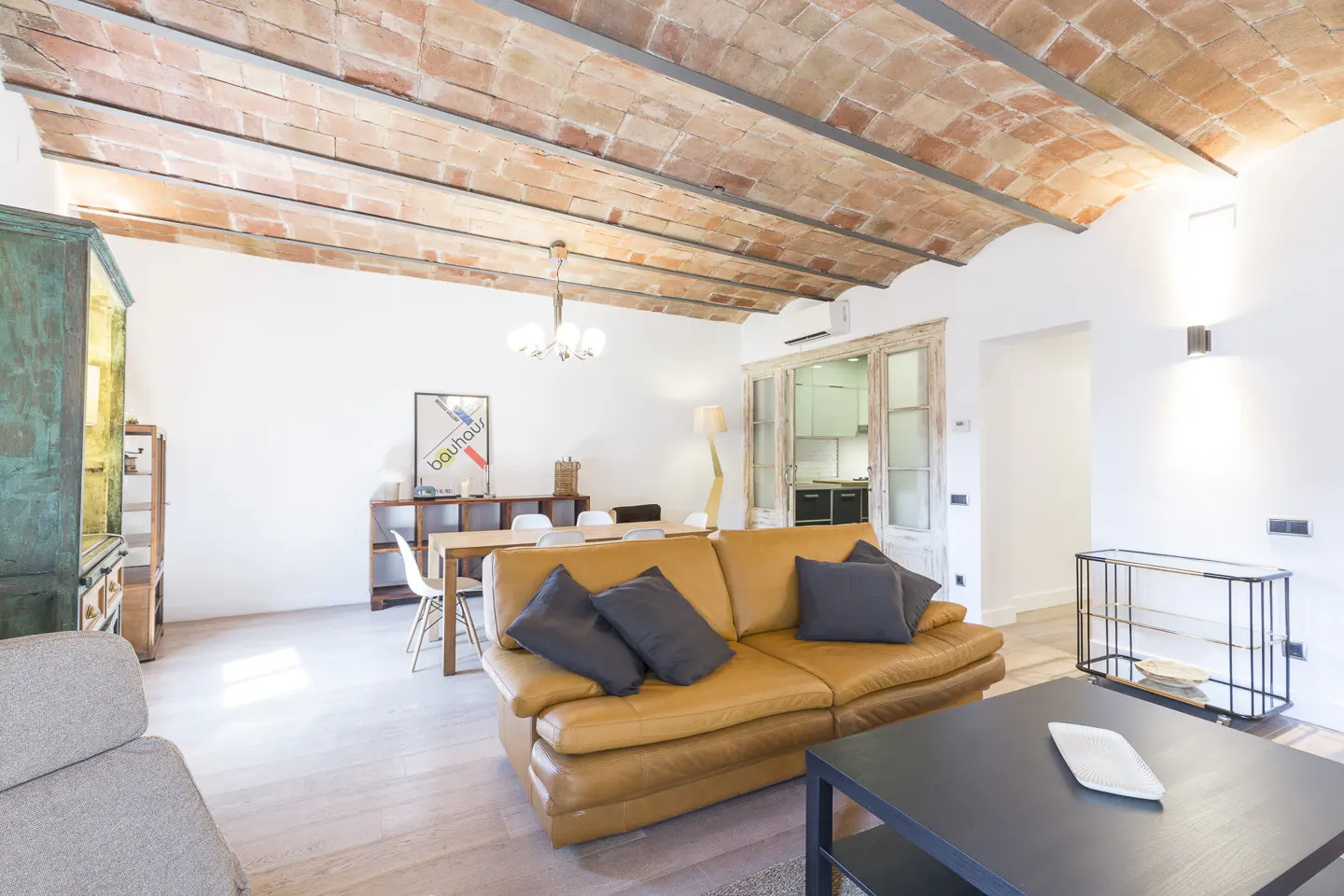 Bright living room with a tan leather sofa, black pillows, and a black coffee table. The ceiling has exposed brick and metal beams.