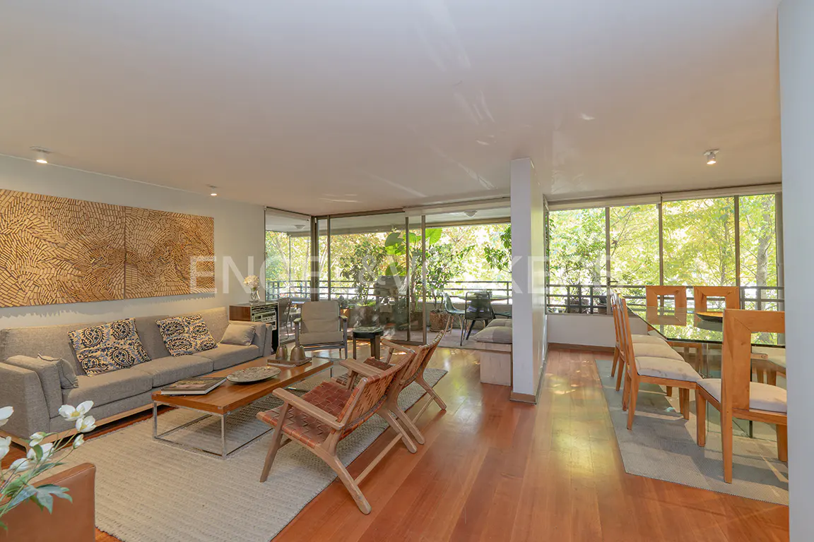 Bright, open-plan living and dining area with wood floors. A gray sofa, wood chairs, and a glass-topped dining table are visible. Balcony with greenery.