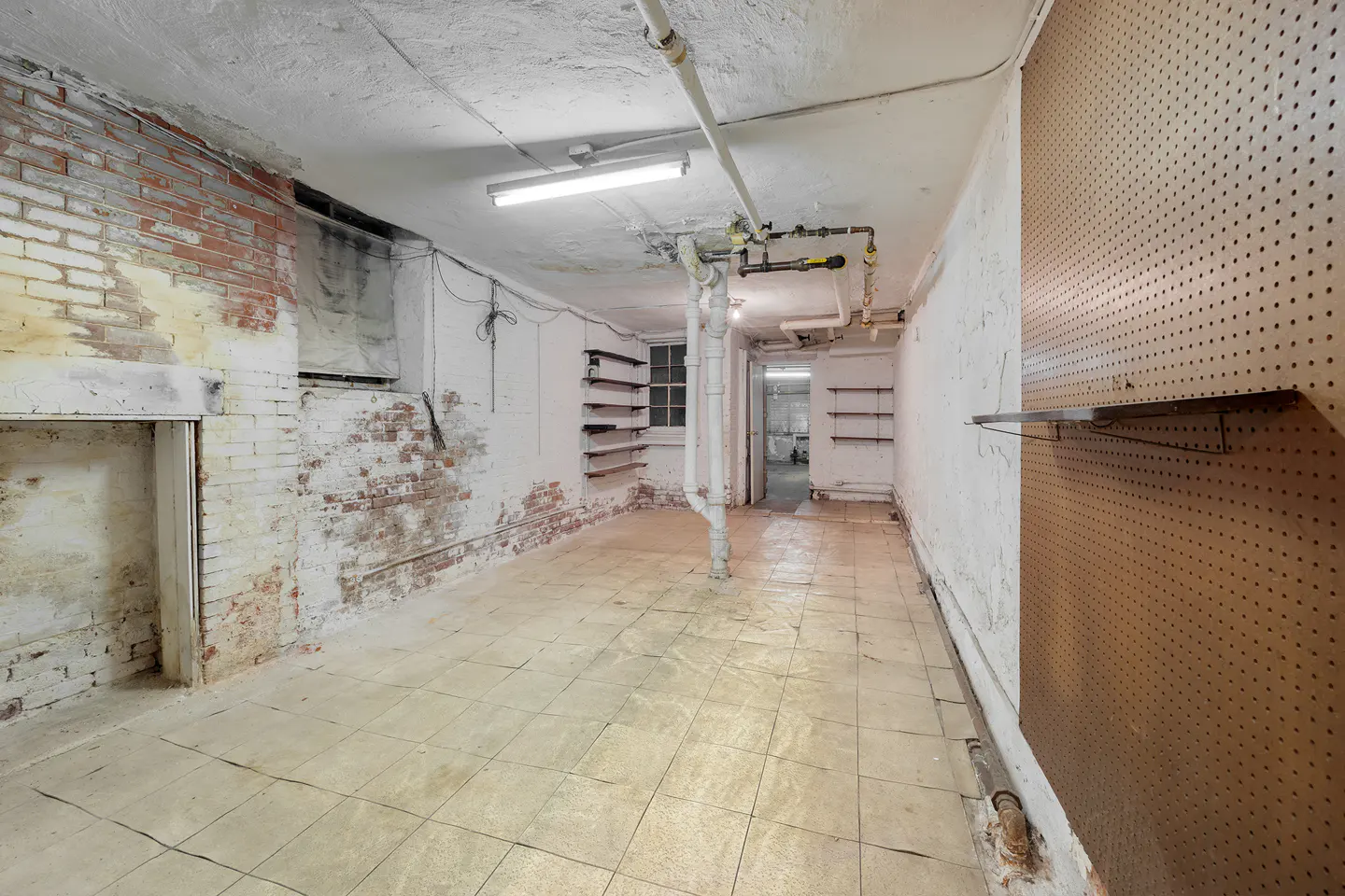 Unfinished basement with exposed brick, white walls, and tiled floor. Pipes run along the ceiling. A pegboard covers part of the right wall.