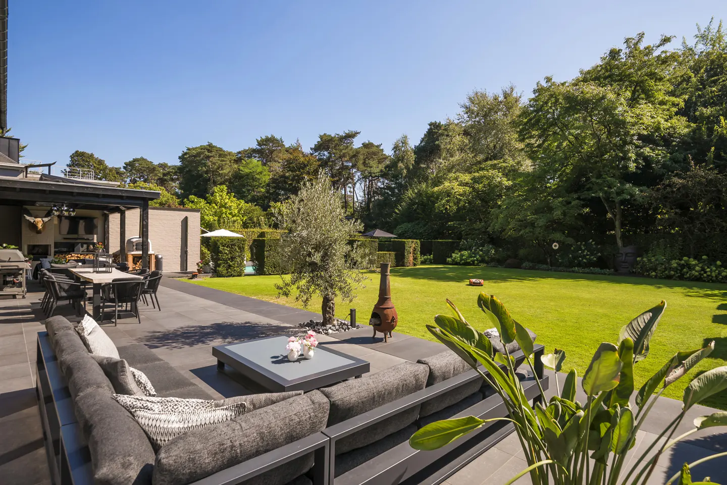 A backyard patio with gray furniture, a table, and a fire pit overlooks a green lawn and trees under a blue sky.