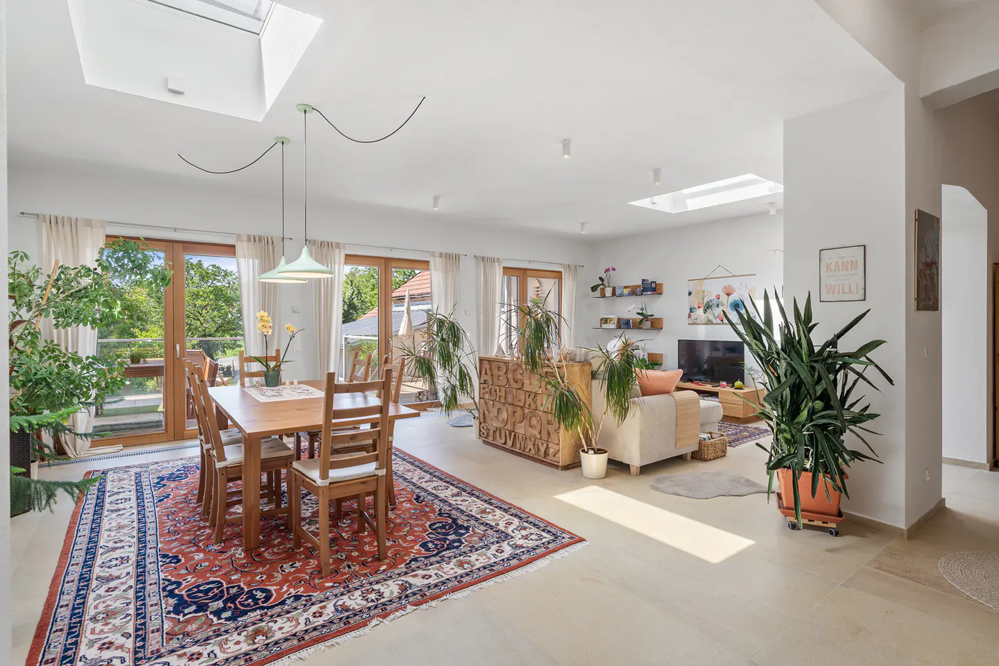 Bright, open-concept living and dining area with wood table, chairs, and a red patterned rug. Large windows and skylights provide natural light.