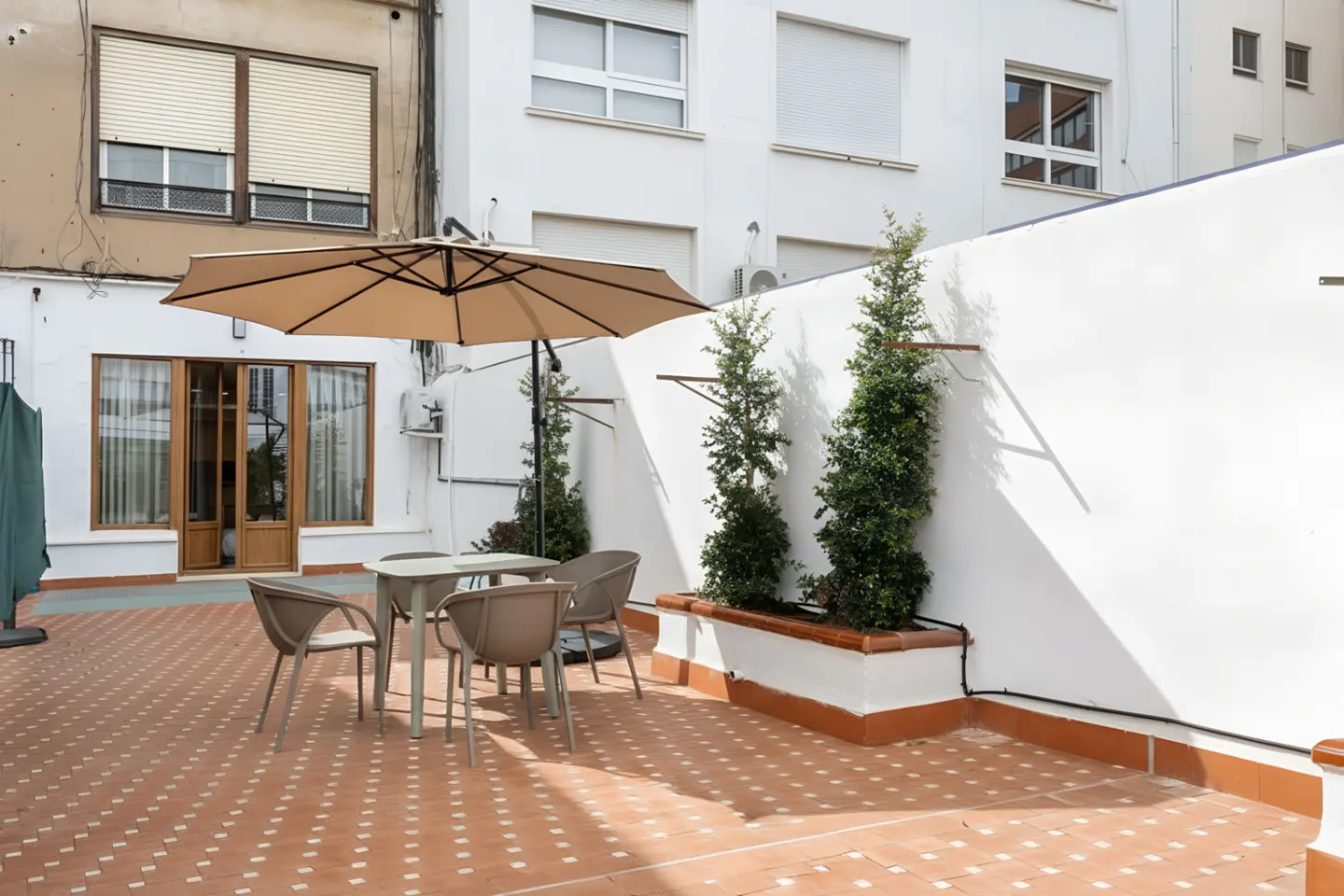Outdoor patio with terracotta tile, table and chairs under a tan umbrella. White walls and green plants add to the ambiance.