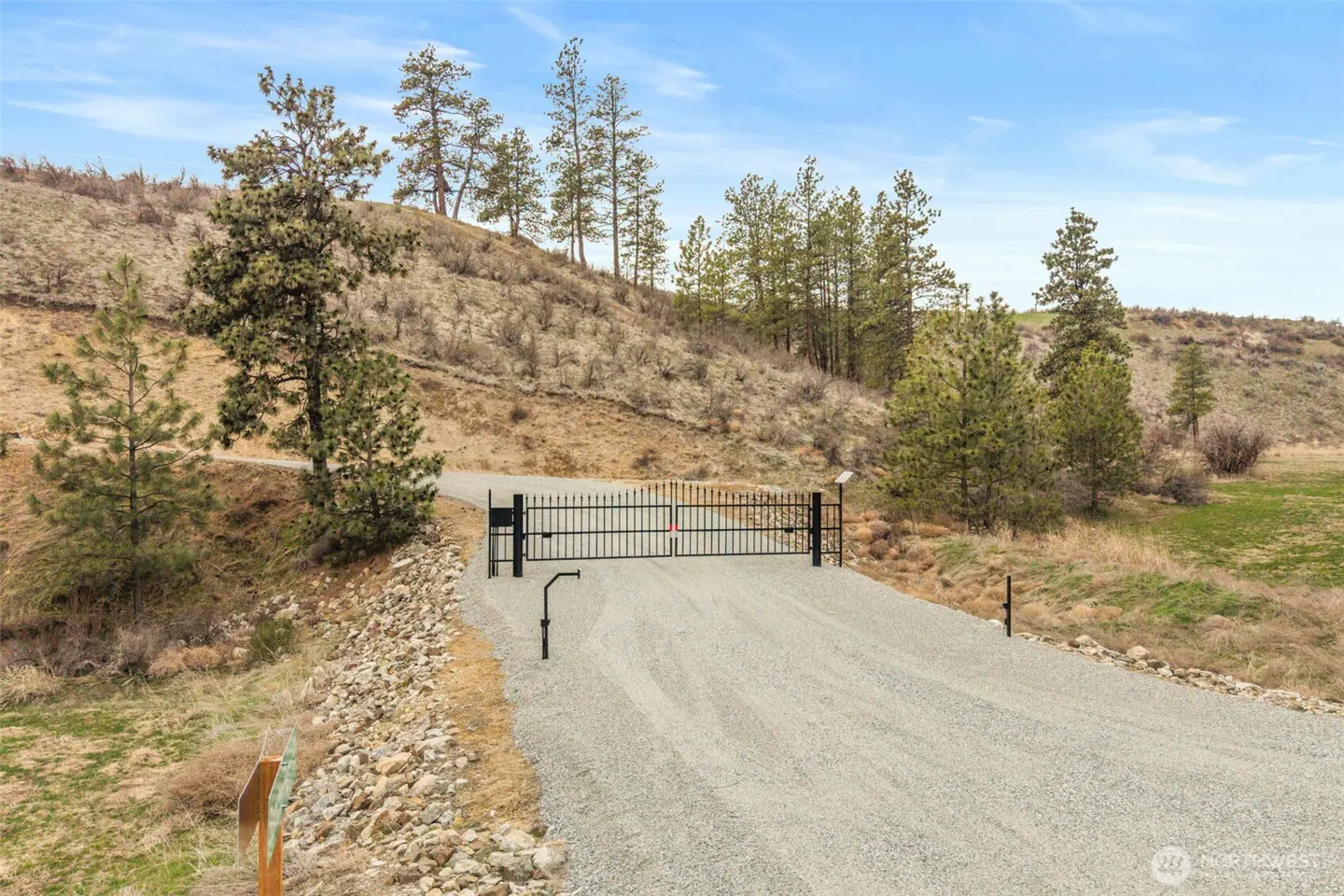 Gated gravel driveway leading up a hill. Trees dot the landscape under a blue sky with light clouds.