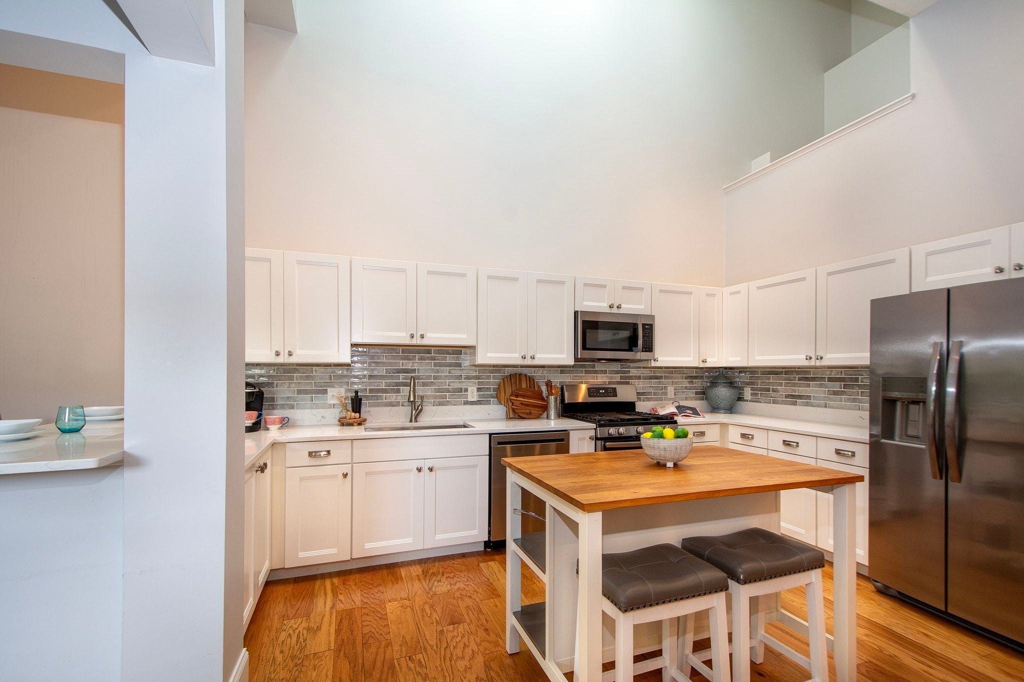 Modern kitchen with white cabinets, stainless steel appliances, a wood-topped island with two stools, and high vaulted ceilings. [AI-generated text]