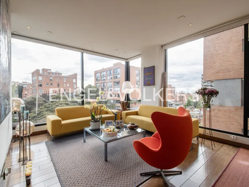 Bright living room with yellow sofas, orange egg chair, and city views through large windows.