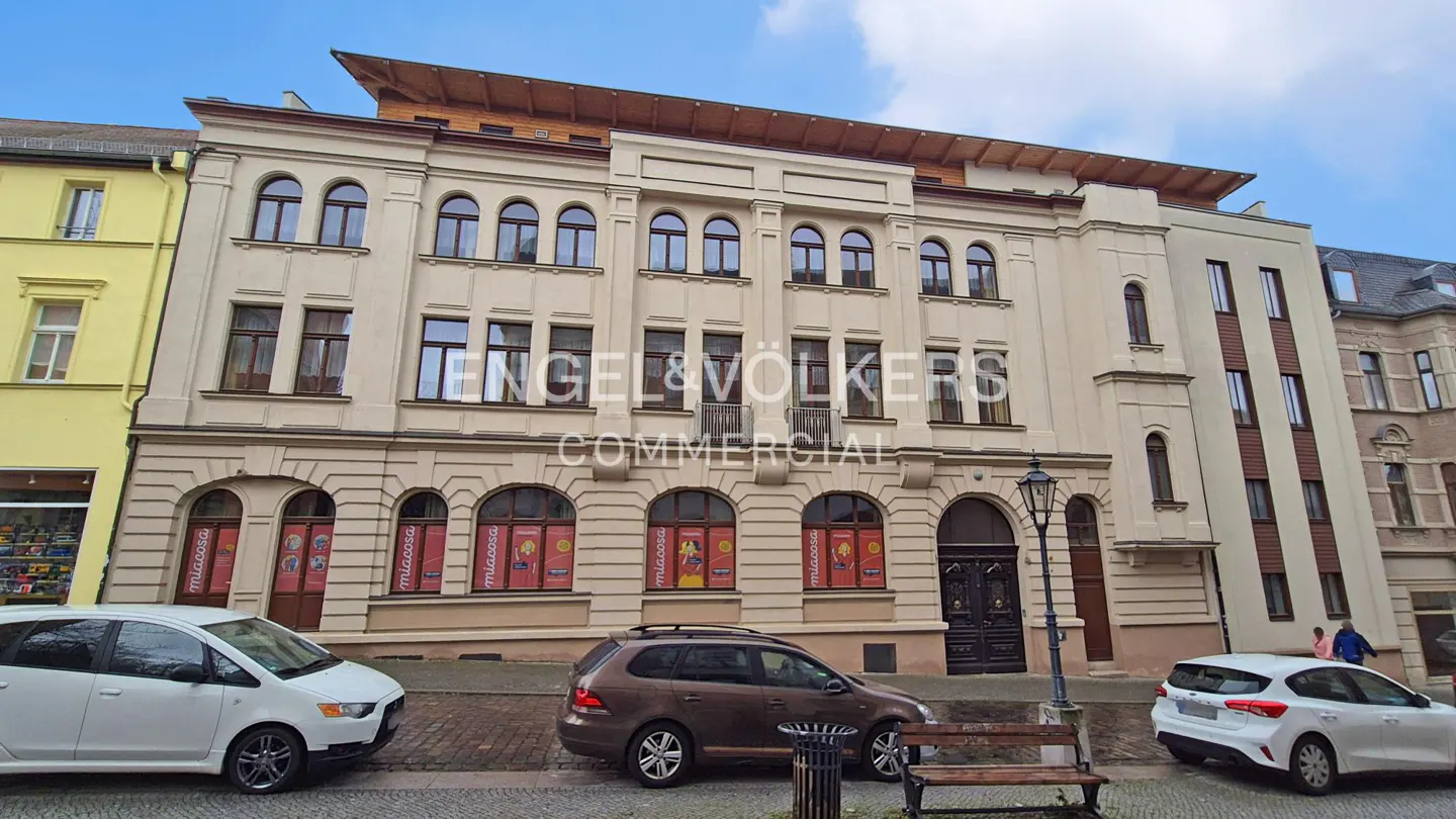 Exterior view of a three-story beige commercial building with arched windows and the Engel & Völkers Commercial logo. Cars are parked in front.