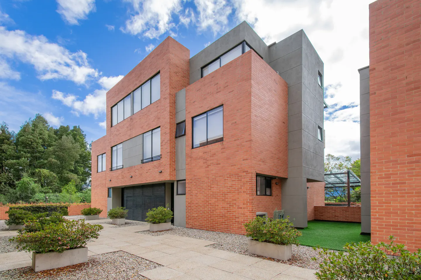 Modern brick and gray concrete building with large windows under a blue sky. Potted plants line the stone walkway.