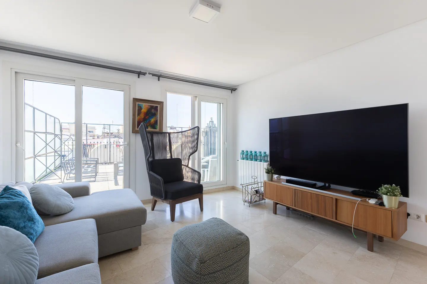 Bright living room with a gray sectional sofa, a tall black chair, and a large TV on a wooden console. Sliding glass doors lead to a balcony.
