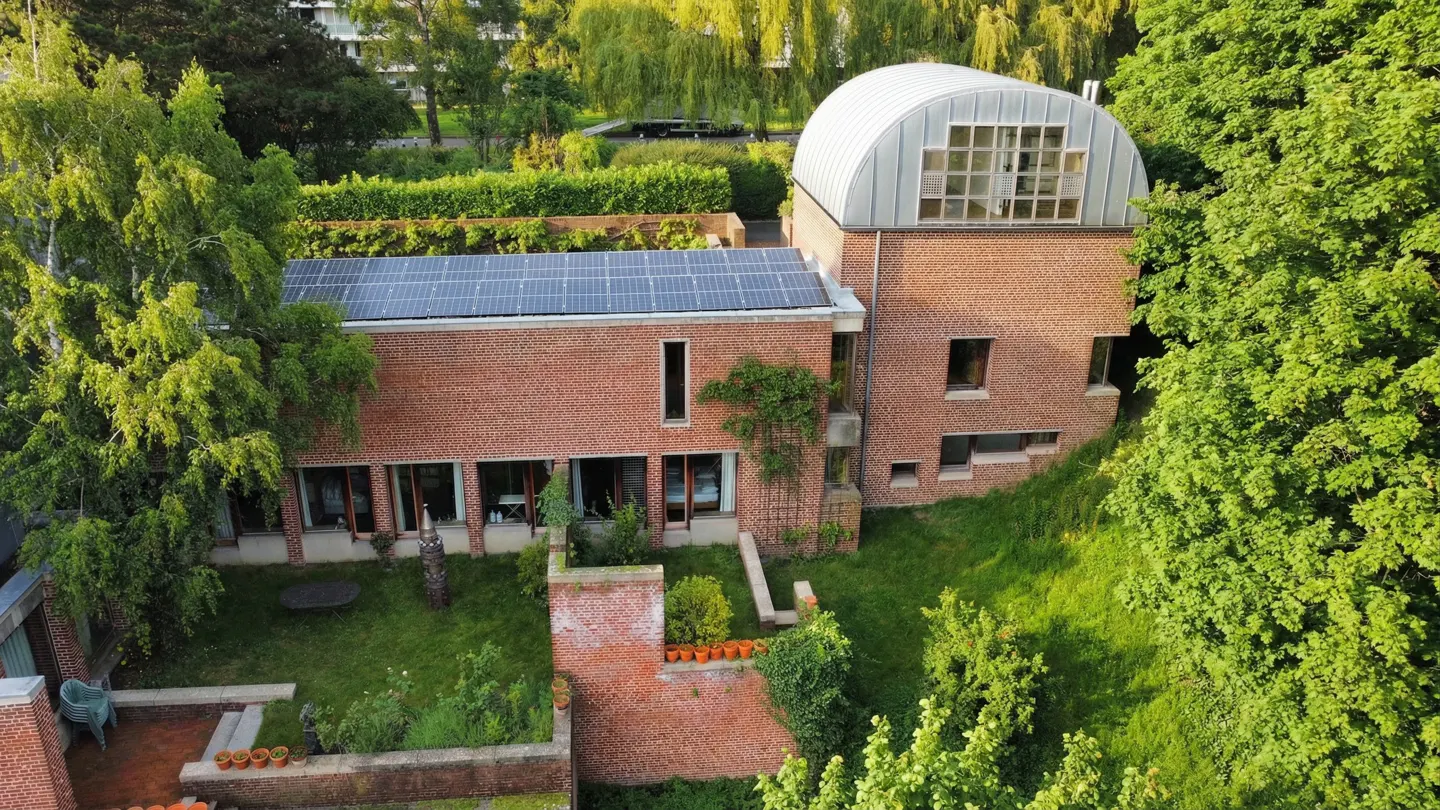 Aerial view of a red brick house with solar panels, a curved roof extension, and a lush green garden.