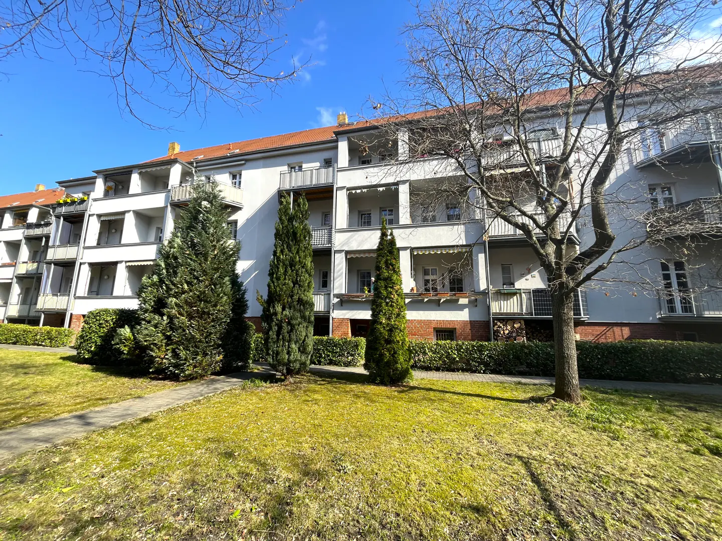 Exterior view of a white apartment building with balconies, a red tile roof, and green lawn with trees.