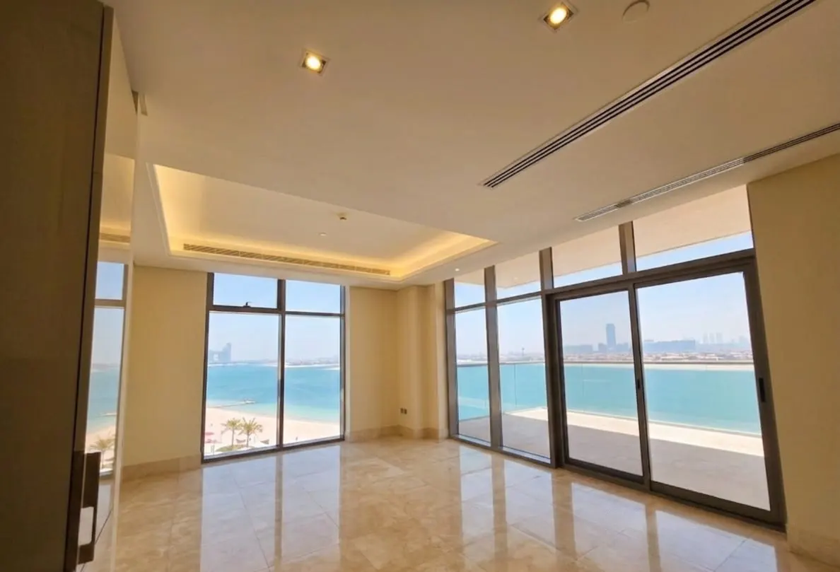 Modern, empty room with beige marble floors, recessed lighting, and floor-to-ceiling windows overlooking a beach and sea.
