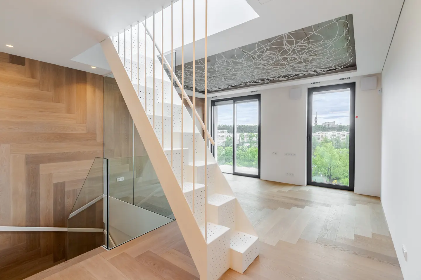 Modern interior with white stairs, glass railing, and wood floors. Large windows offer a city view. Ceiling has a unique, artistic design.