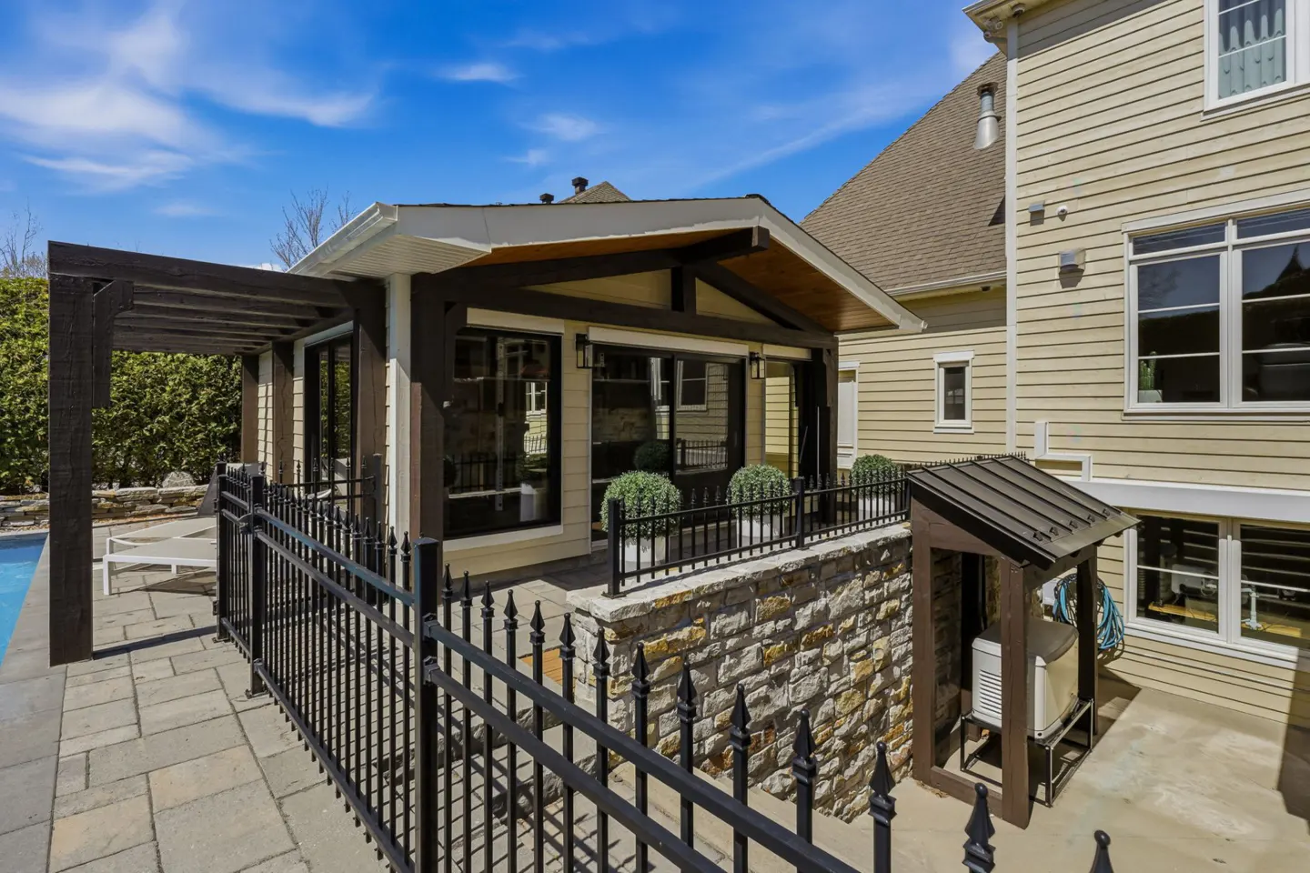 Exterior view of a tan house with a dark wood pergola, stone wall, and black iron fence. A pool is visible on the left.