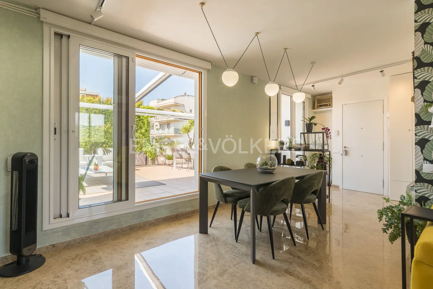 A bright dining room with a dark table and green chairs, leading to a sunny terrace with outdoor furniture.