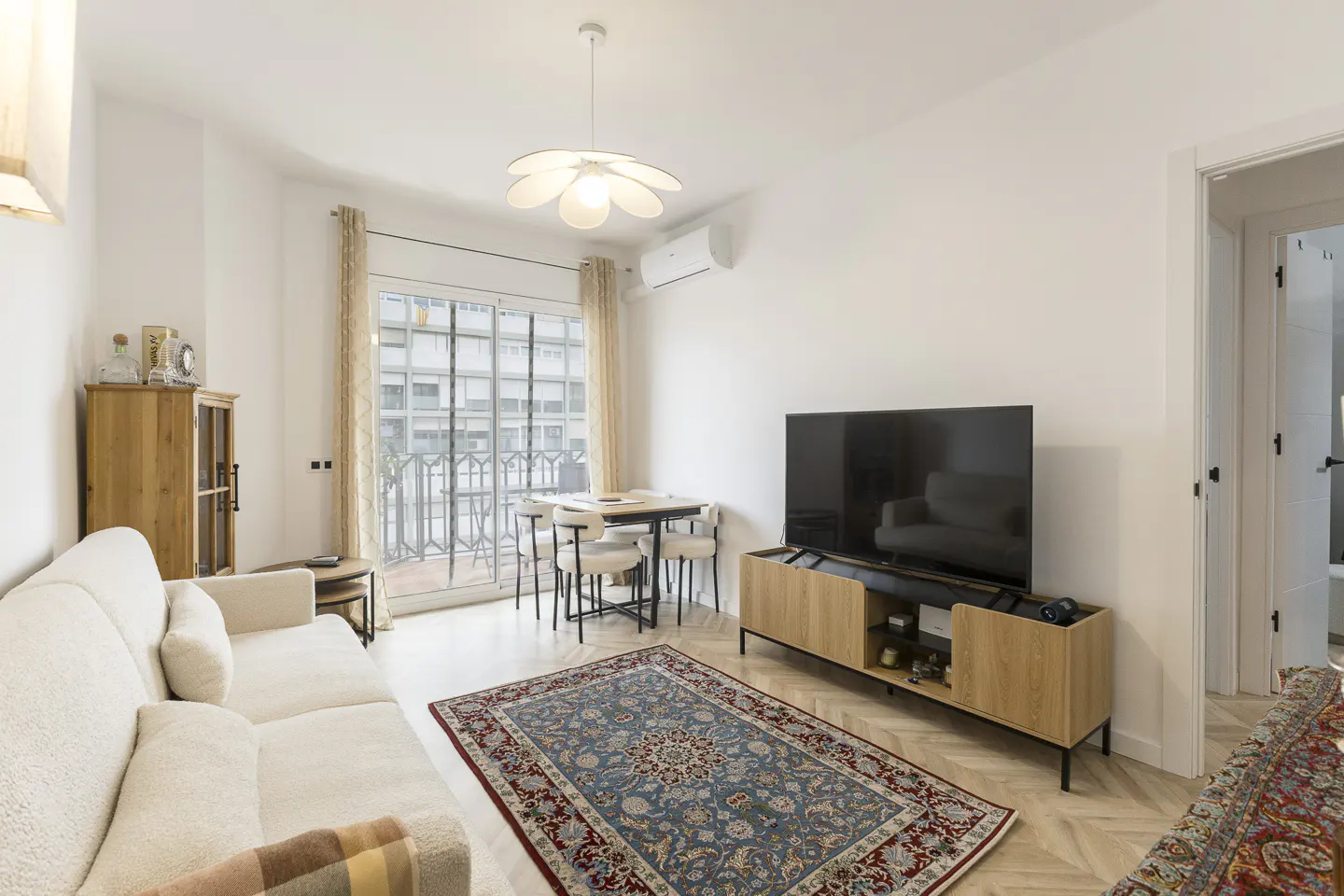 Bright living room with white walls, a cream sofa, a TV on a wooden stand, and a blue and red rug. A dining table sits near a window.